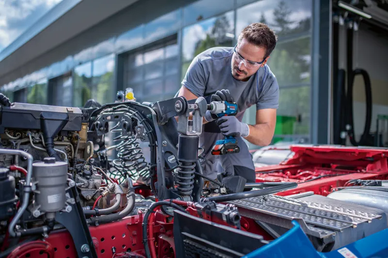 안전 장비를 착용한 사람이 차량 섀시에 무선 충격 렌치를 사용하고 있습니다.