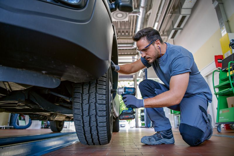 안전 장비를 착용한 사람이 무선 충격 렌치를 사용하여 자동차 바퀴 볼트를 조이고 있습니다.
