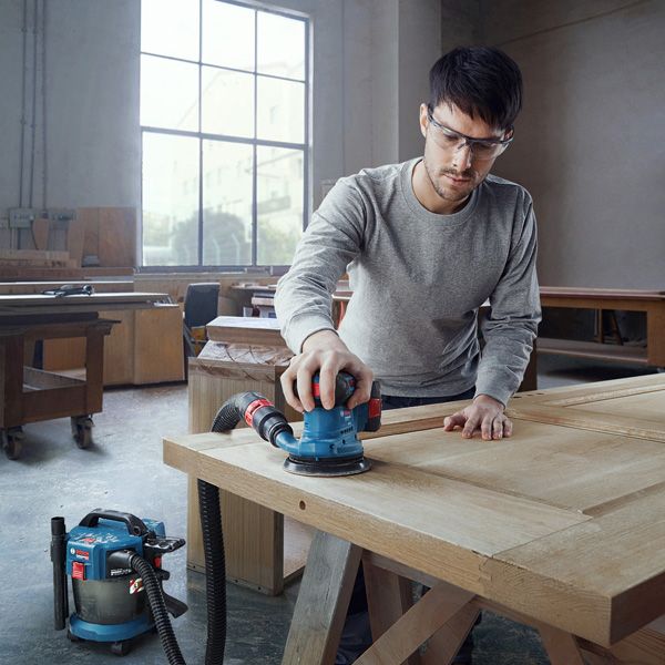 안전장비를 착용한 사람이 궤도 샌더를 이용해 나무 문을 샌딩하고 있습니다.