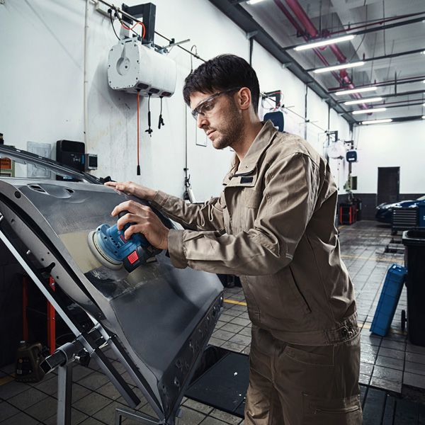 안전장비를 착용한 사람이 전동 샌더로 자동차 문을 샌딩하고 있습니다.