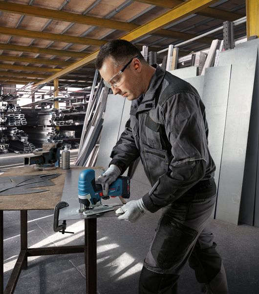 GST 18V-125 S 안전 장비를 착용한 사람이 작업장에서 무선 톱으로 금속판을 자르고 있습니다.