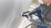 안전 장비를 착용한 사람이 무선 건식벽체 라우터를 사용하여 건식벽체에 구멍을 뚫고 있습니다.