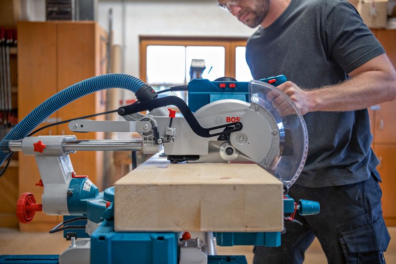 안전 장비를 착용한 사람이 마이터 톱을 조작하여 나무 들보를 자르고 있습니다.