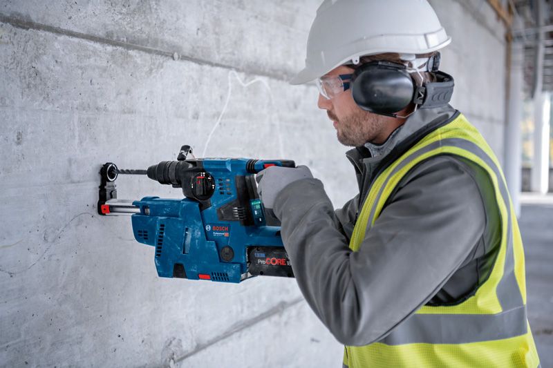 안전장비를 착용한 사람이 회전 망치를 이용해 콘크리트 벽에 구멍을 뚫고 있습니다.