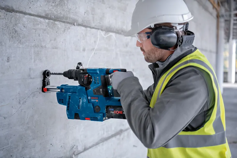 안전장비를 착용한 사람이 회전 망치를 이용해 콘크리트 벽에 구멍을 뚫고 있습니다.