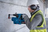 안전장비를 착용한 사람이 회전 망치를 이용해 콘크리트 벽에 구멍을 뚫고 있습니다.