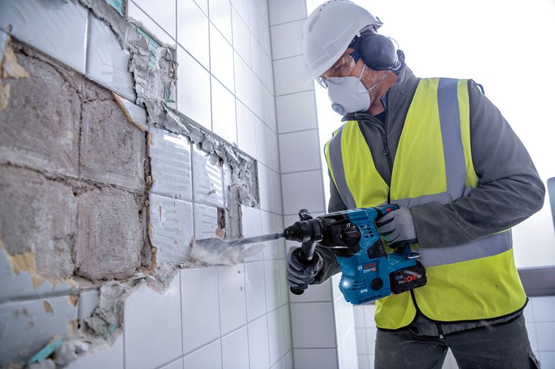 안전장비를 착용한 사람이 회전 망치를 이용해 벽에서 타일을 제거하고 있습니다.