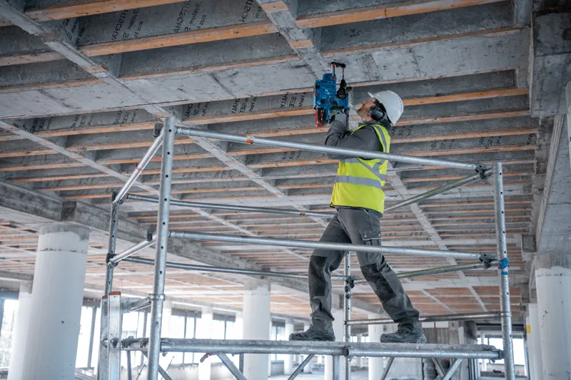 안전장비를 착용한 사람이 비계 위에 서서 콘크리트 천장에 코어 드릴을 사용하고 있습니다.