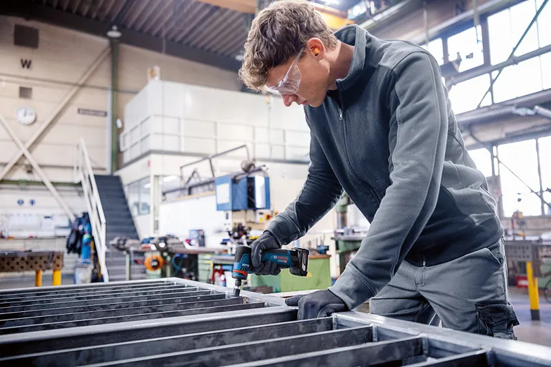 안전 장비를 착용한 사람이 작업장에서 전동 공구를 사용해 금속 보를 고정하고 있습니다.