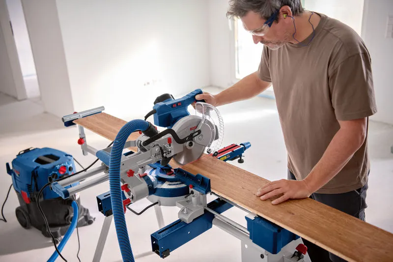 안전장비를 착용한 사람이 마이터 톱으로 나무판을 자르고 있습니다.