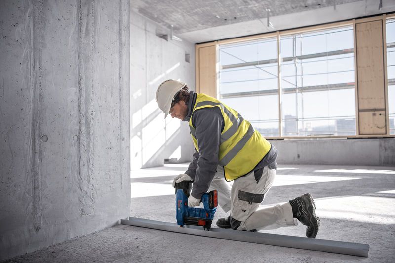 안전장비를 착용한 사람이 무선 콘크리트 못 박는 기계를 사용해 금속 트랙을 바닥에 고정하고 있습니다.