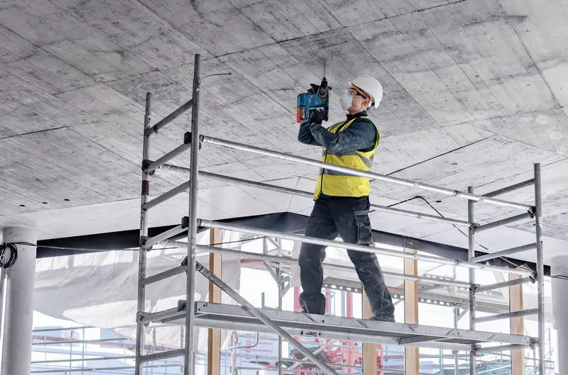 안전장비를 착용한 사람이 비계 위에 서서 콘크리트 천장에 구멍을 뚫고 있습니다.