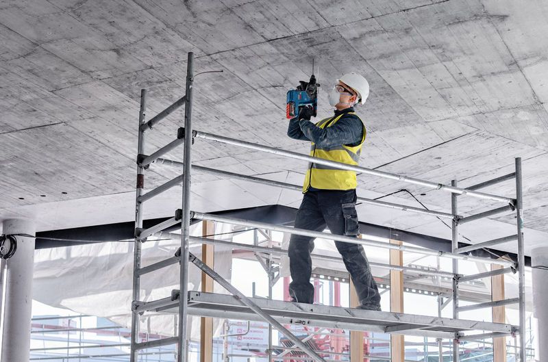 안전장비를 착용한 사람이 비계 위에 서서 콘크리트 천장에 구멍을 뚫고 있습니다.