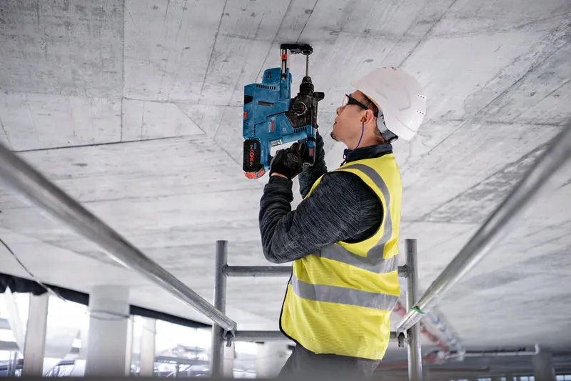 안전장비를 착용한 사람이 전동공구를 이용해 콘크리트 천장에 구멍을 뚫고 있습니다.