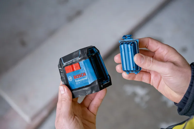 한 사람이 실내에서 파란색 레이저 레벨링 도구와 배터리 칸을 잡고 있습니다.