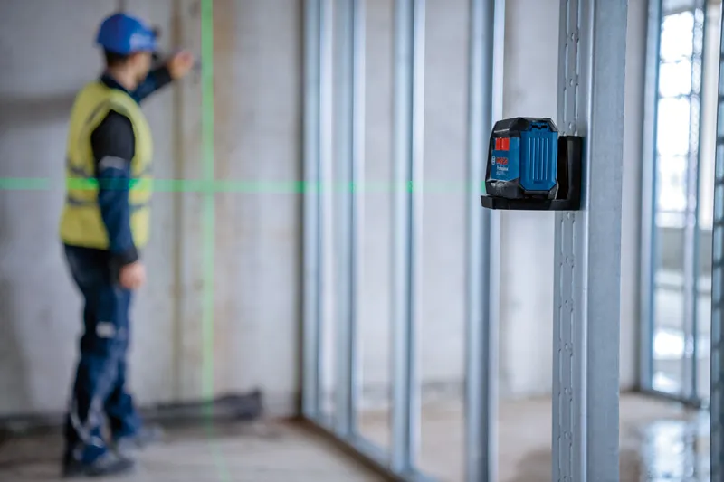 안전 장비를 착용한 사람이 레이저 레벨링 도구를 사용하여 벽 프레임을 정렬하고 있습니다.