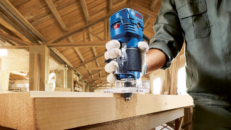 안전 장비를 착용한 사람이 작업장에서 휴대용 라우터를 이용해 나무를 다듬고 있습니다.
