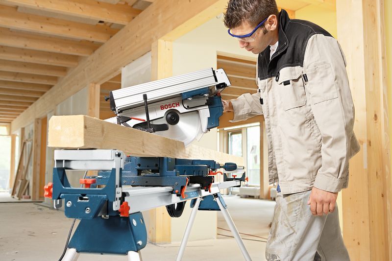 안전 장비를 착용한 사람이 마이터 톱을 사용하여 실내에서 나무 들보를 자르고 있습니다.