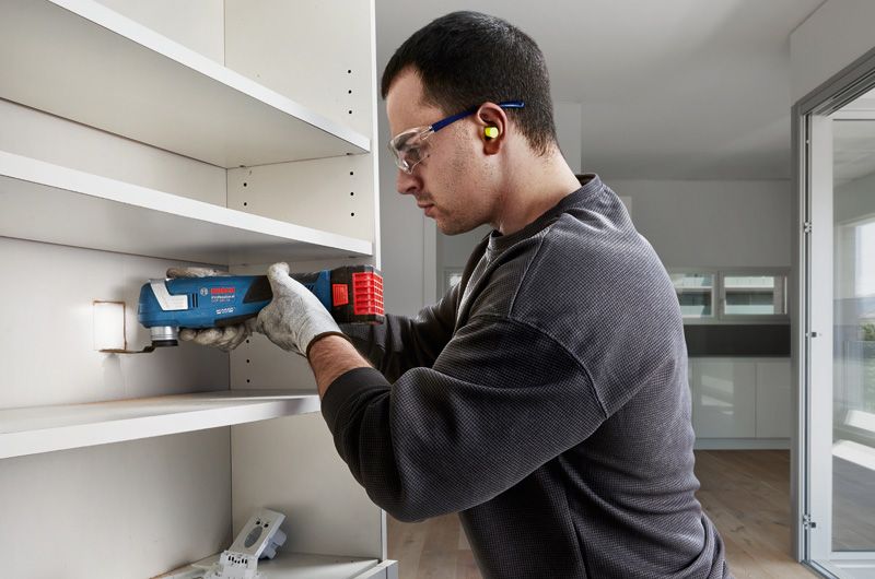 안전 장비를 착용한 사람이 멀티툴을 이용해 선반 패널을 자르고 있습니다.