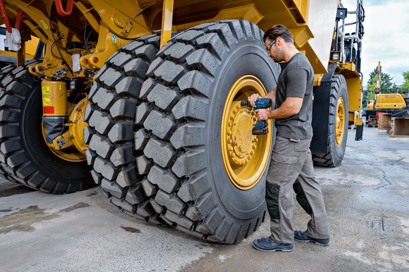 안전장비를 착용한 사람이 대형 건설 차량 타이어에 임팩트 렌치를 사용하고 있습니다.
