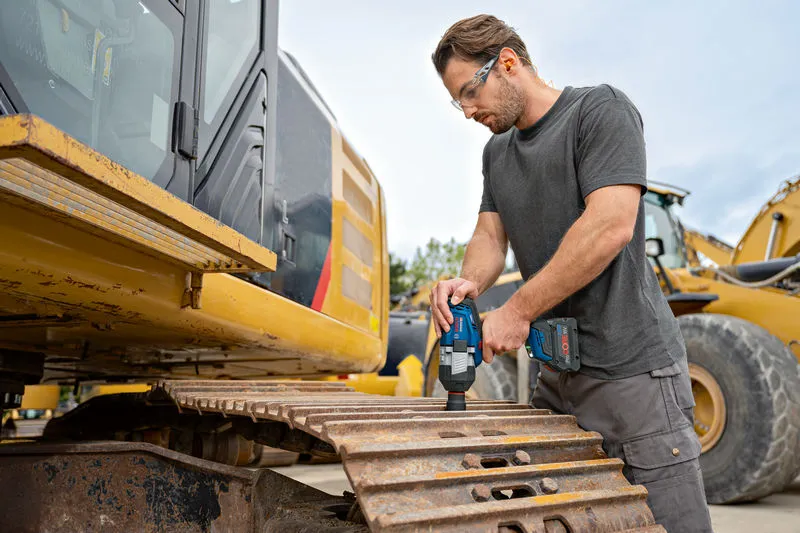 안전 장비를 착용한 사람이 굴삭기 트랙에서 무선 충격 렌치를 사용하고 있습니다.