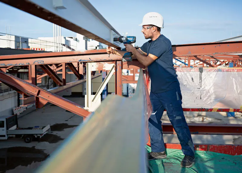 안전 장비를 착용한 사람이 무선 충격 렌치를 사용하여 강철 보를 작업하고 있습니다.