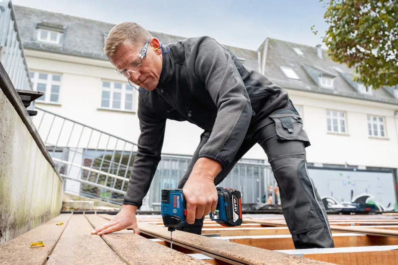 안전장비를 착용한 사람이 무선 드릴을 사용해 데크 보드를 고정하고 있습니다.