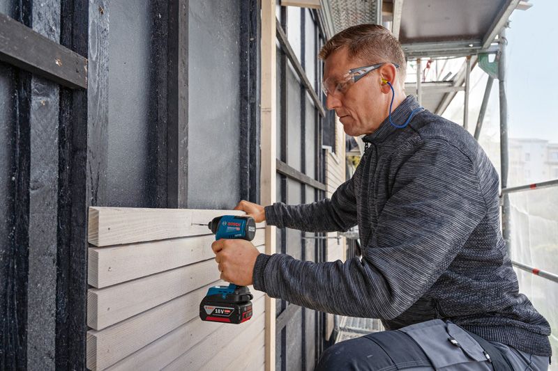 안전장비를 착용한 사람이 무선 드릴을 이용해 나무 패널을 벽에 고정하고 있습니다.