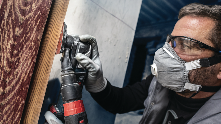 안전 장비를 착용한 사람이 전동 공구로 나무 표면을 연마하고 있습니다.