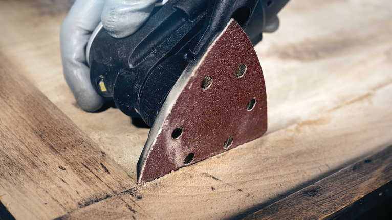 안전 장비를 착용한 사람이 디테일 샌더를 이용해 나무 표면을 샌딩하고 있습니다.