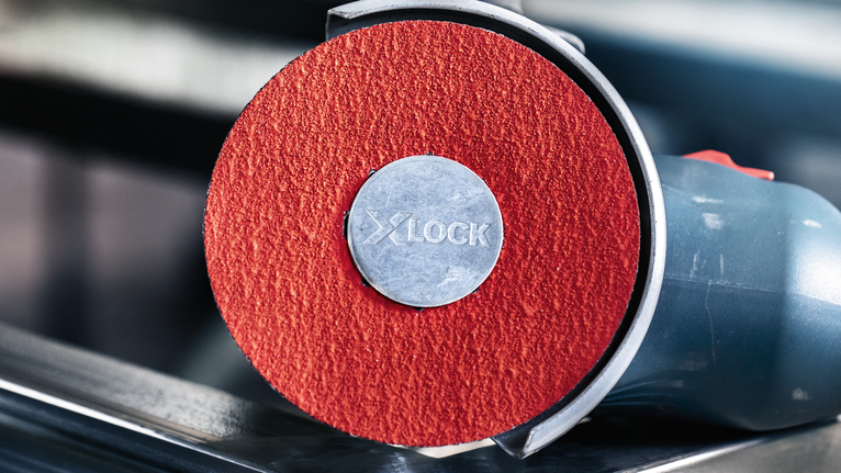 PRO R780 X-Lock 화이버 디스크, 125mm, G36 금속 위에 놓인 붉은 연마 디스크가 있는 각도 분쇄기의 클로즈업.
