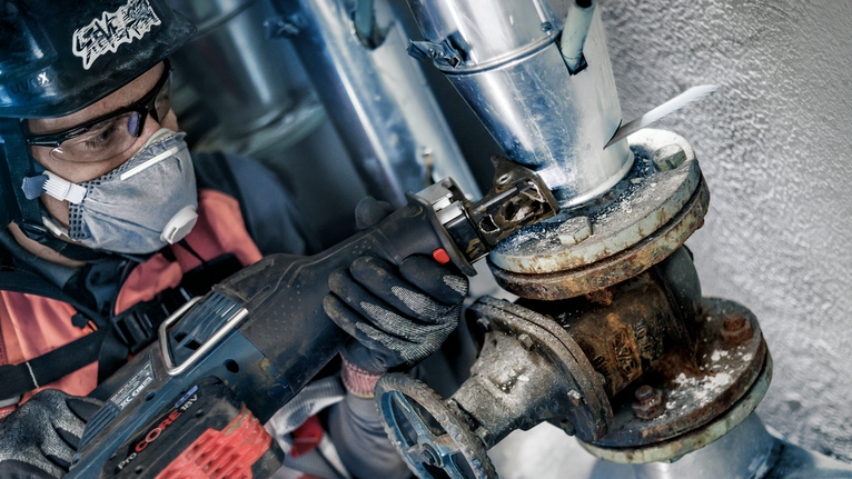 안전 장비를 착용한 사람이 왕복톱을 사용해 부식된 파이프를 절단하고 있습니다.