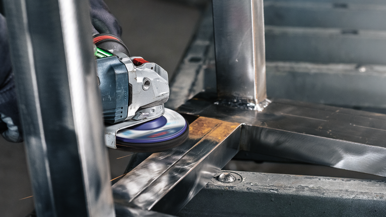 PRO N377 클리닝 디스크, 125mm, 22.23mm, X-Lock 안전 장비를 착용한 사람이 각도 그라인더로 금속 프레임을 연마하고 있습니다.