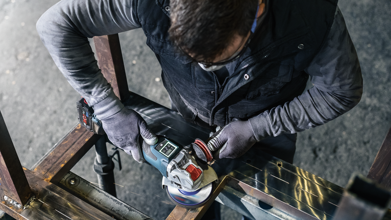 PRO N377 클리닝 디스크, 125mm, 22.23mm, X-Lock 안전 장비를 착용한 사람이 각도 분쇄기로 금속 빔을 갈고 있습니다.