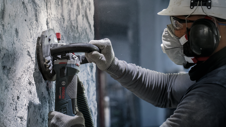 안전장비를 착용한 사람이 전동공구를 이용해 콘크리트 벽을 갈고 있습니다.