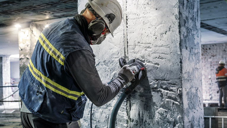 안전 장비를 착용한 사람이 전동 공구로 콘크리트 기둥을 갈아내고 있습니다.