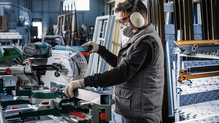 안전 장비를 착용한 사람이 마이터 톱을 조작하여 금속 프로필을 절단하고 있습니다.