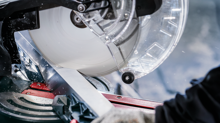 안전 장비를 착용한 사람이 마이터 톱을 사용하여 금속 조각을 자르고 있습니다.