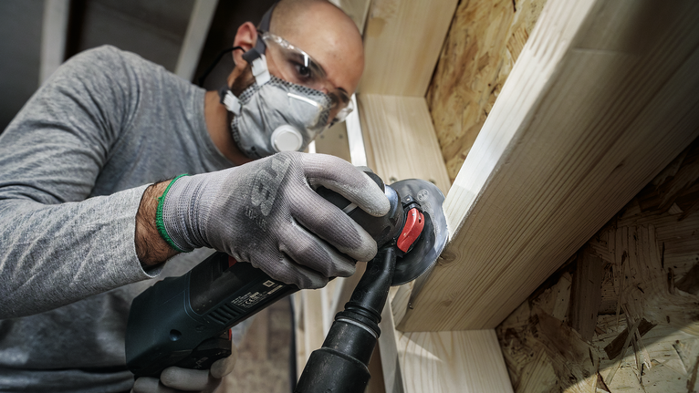 안전 장비를 착용한 사람이 각도 그라인더를 사용하여 나무 프레임을 연마하고 있습니다.