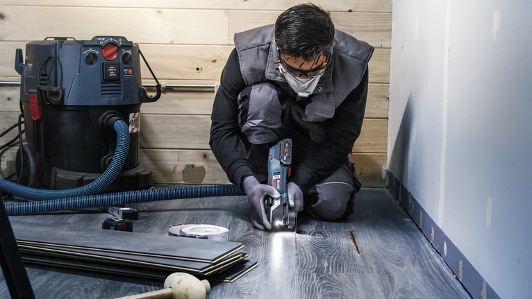 안전장비를 착용한 사람이 전동공구를 이용해 라미네이트 바닥을 자르고 있습니다.