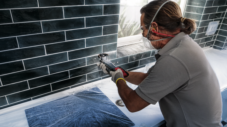 안전 장비를 착용한 사람이 전동 공구를 사용하여 창문 아래의 타일을 자르고 있습니다.