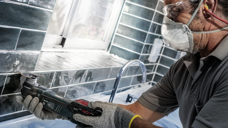 안전 장비를 착용한 사람이 전동 공구를 사용하여 싱크대 근처의 타일을 자르고 있습니다.