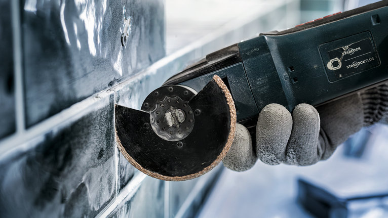 PRO ACZ 85 RT3 멀티커터 날, 85 x 2.5mm 안전 장비를 착용한 사람이 전동 공구를 사용하여 타일을 자르고 있습니다.