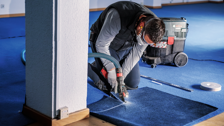 PRO ASZ 32 SC 멀티커터 날, 24 x 11mm 안전 장비를 착용한 사람이 실내에서 전동 공구를 이용해 파란색 카펫을 자르고 있습니다.