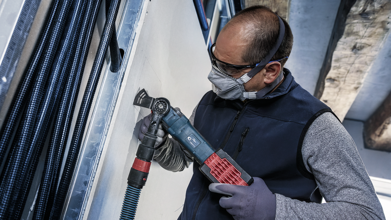 안전장비를 착용한 사람이 전동공구를 이용해 건식벽체를 자르고 있습니다.