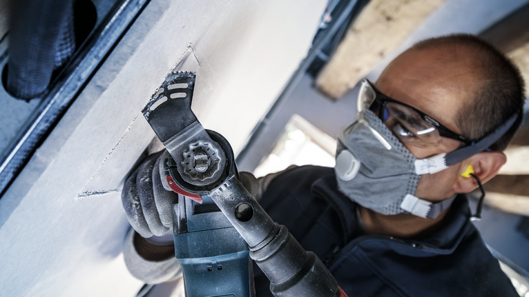 안전장비를 착용한 사람이 전동공구를 이용해 건식벽체를 자르고 있습니다.