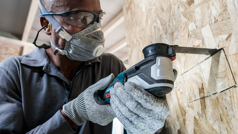 안전 장비를 착용한 사람이 전동 공구를 사용해 나무에 정사각형 구멍을 자릅니다.