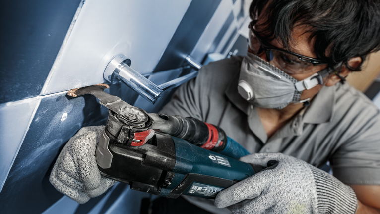 PRO MATI 68 RT3 멀티커터 날, 2.8 x 68mm 안전 장비를 착용한 사람이 전동 공구를 사용하여 파이프 주변의 타일을 자르고 있습니다.