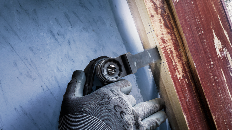 안전 장비를 착용한 사람이 멀티툴을 사용하여 페인트칠된 표면 근처의 나무를 자릅니다.