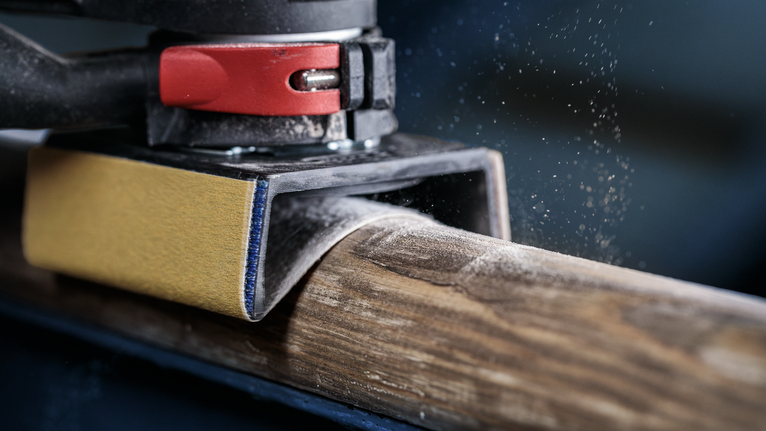 PRO AUZ 70 G 프로파일 샌더, 70 x 65mm, 5개 궤도 샌더로 둥근 나무 들보를 매끄럽게 다듬고 있는데, 나무 먼지 입자가 보입니다.